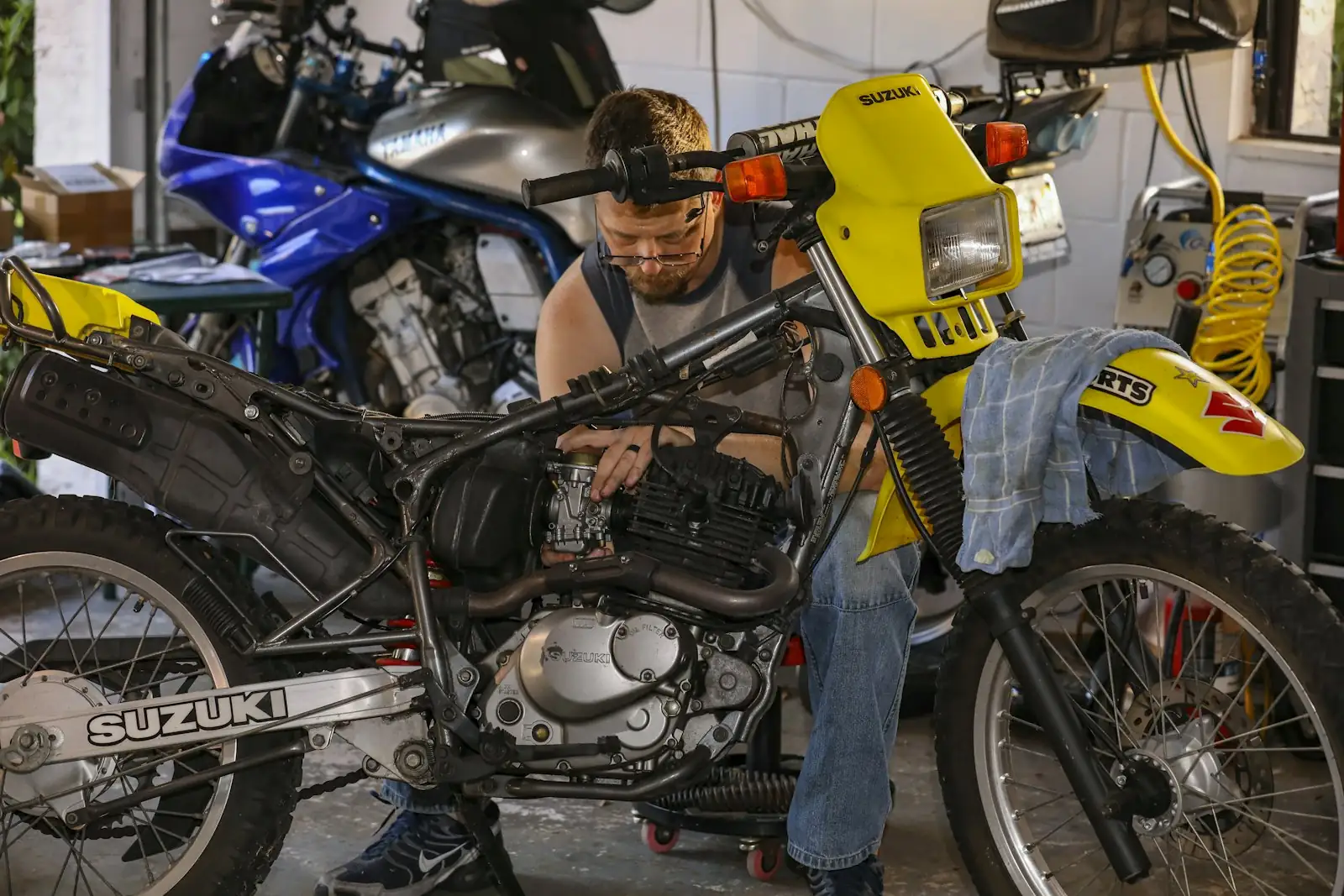 A rider working on the engine of a yellow Suzuki dirt bike in his home garage