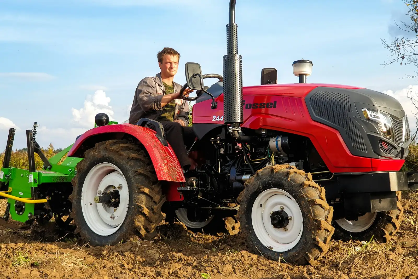 A farmer driving a small red tractor with a rotary hoe across a ploughed field