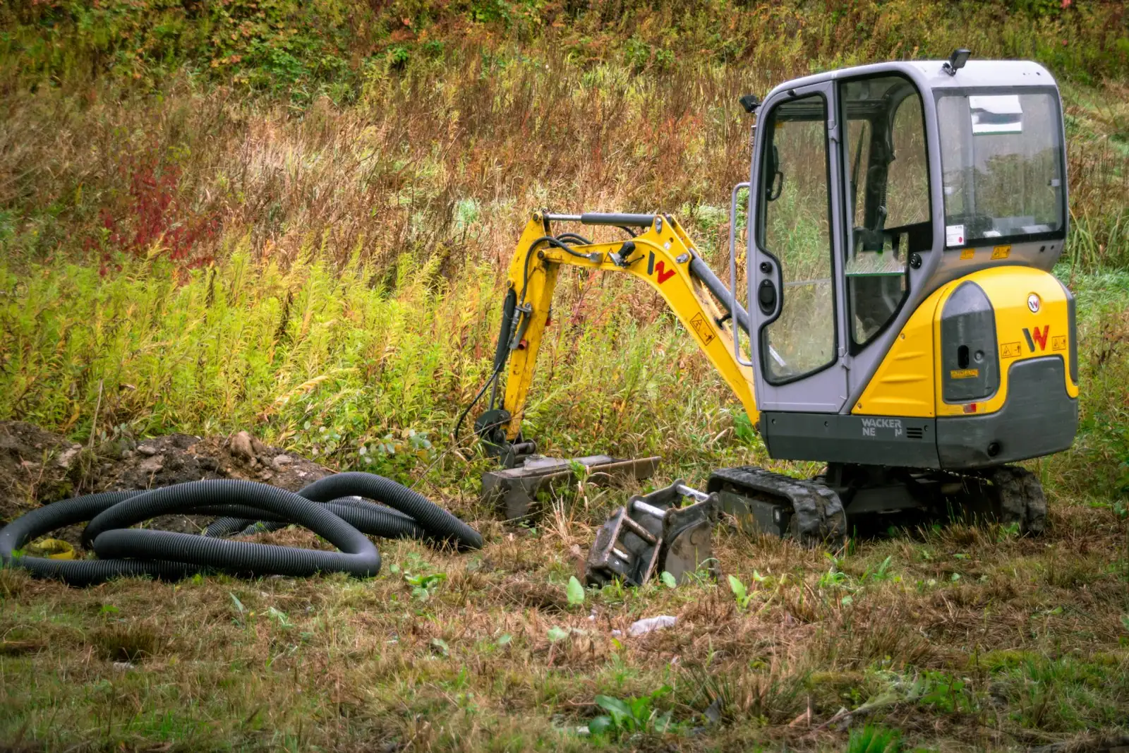 A yellow mini excavator on rough ground beside coiled drainage pipe